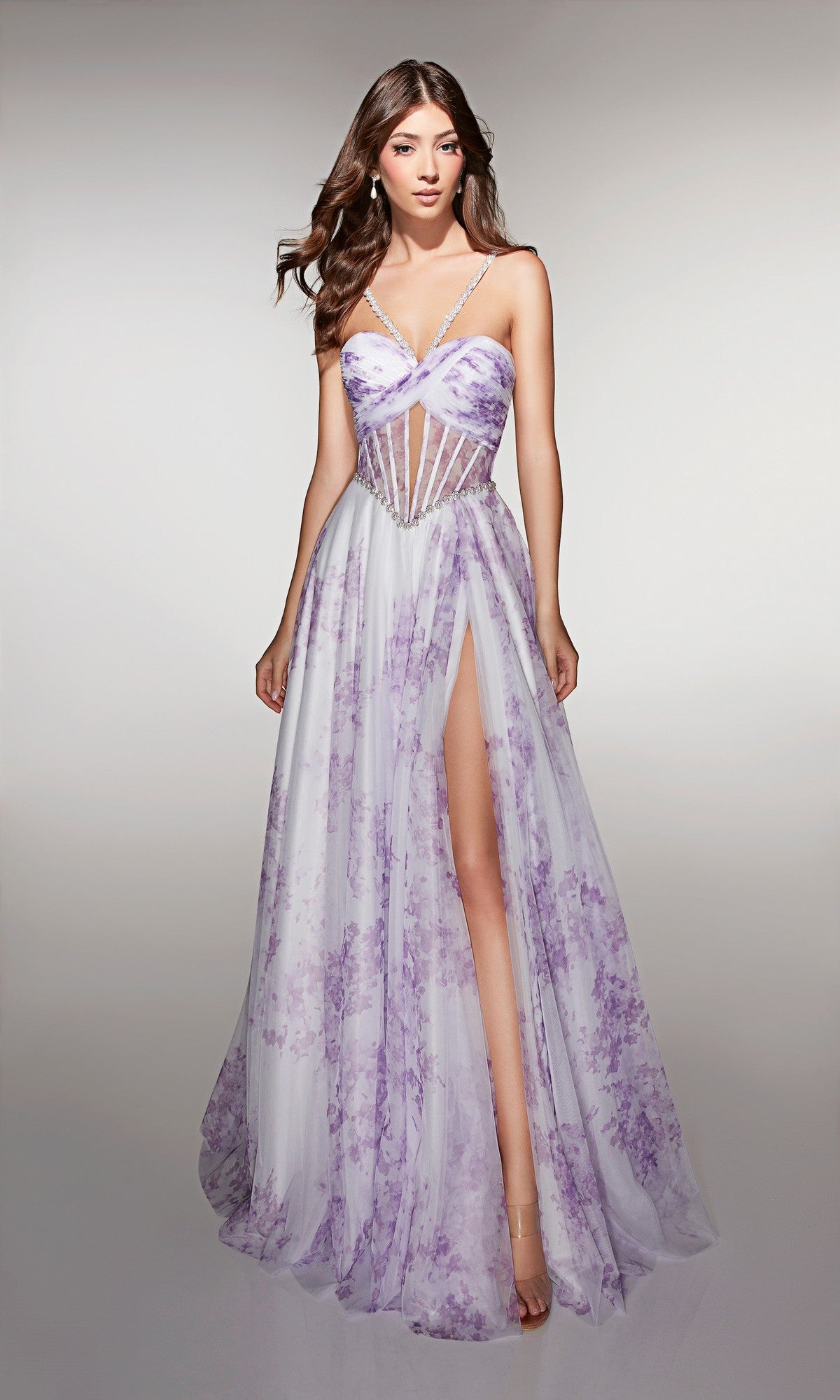 A woman stands against a plain background wearing the Alyce 61994 long prom dress featuring a sleeveless floral design, sheer corset bodice, thin straps, and a high slit.
